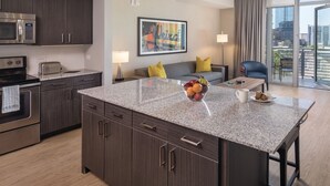 Una cocina moderna con armarios de madera oscura, encimera de granito y electrodomésticos de acero inoxidable. Cuenta con una zona de comedor con mesa y sillas, un sofá con cojines amarillos y un amplio ventanal con vistas a la ciudad.