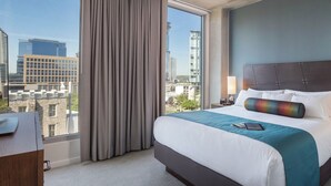 Habitación de hotel con una cama grande, un escritorio, una silla y vistas a la ciudad a través de amplios ventanales.