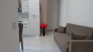 En moderne stue med en beige sofa og et lille bord. Der er et køkkenområde med hvide skabe og en komfur. En vase med røde blomster står på gulvet nær sofaen.