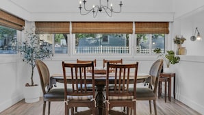Un comedor con una mesa redonda, sillas de madera, una araña y ventanas grandes con persianas de bambú.