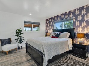 Un dormitorio con cama, mesitas de noche, una silla, una planta en maceta y una ventana.
