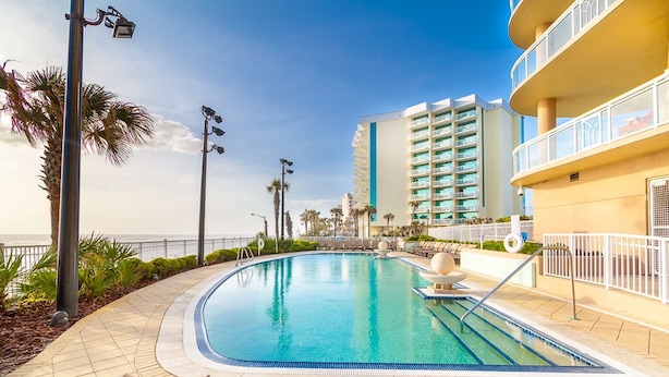 A swimming pool with lounge chairs, a palm tree, and a high-rise building in the background. - Fabulous direct ocean front with private balcony & steps to the beach (Daytona Beach)