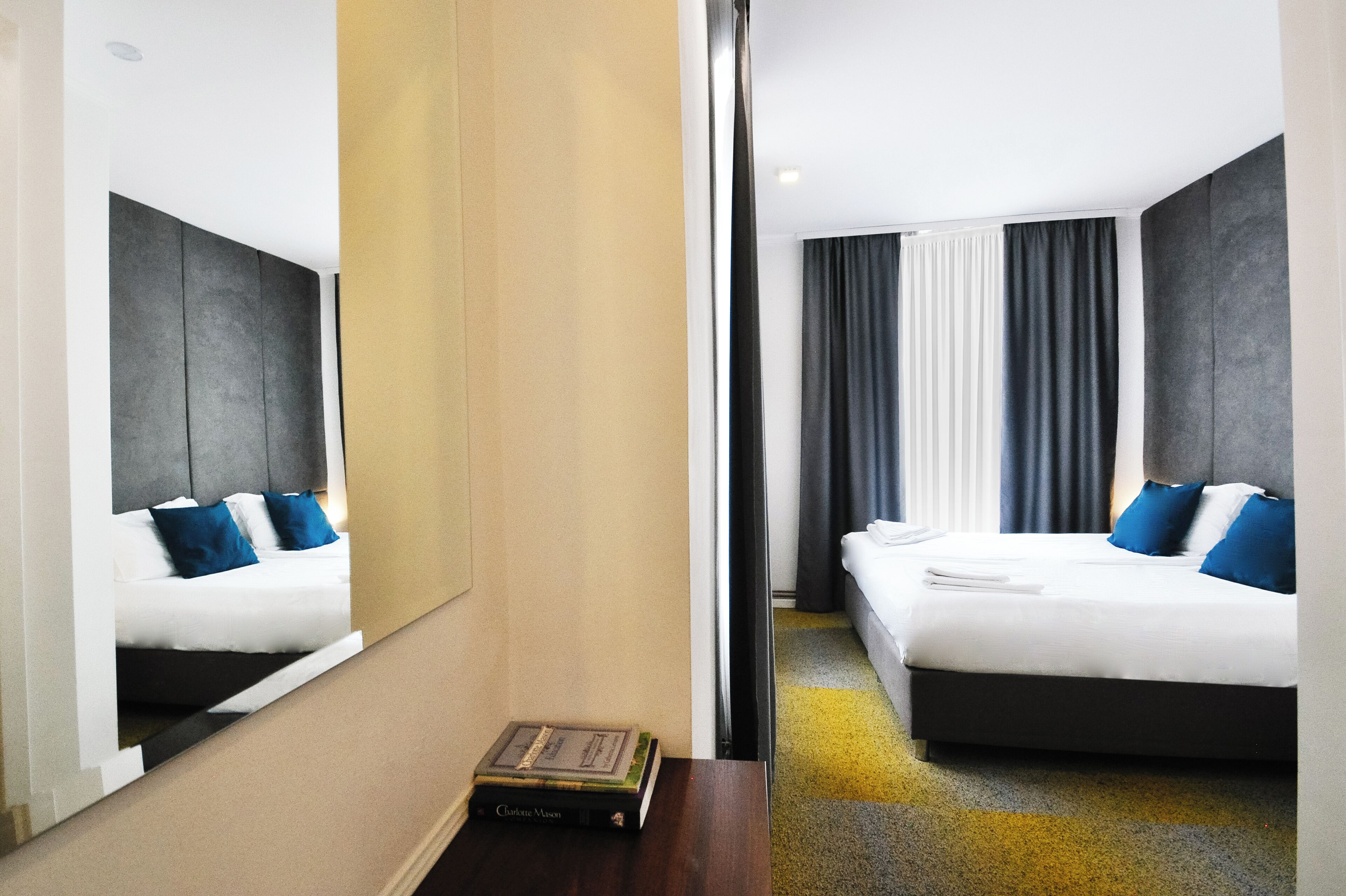 Ein Hotelzimmer mit einem großen Bett, einem Nachttisch mit einem Buch, einem Spiegel und Blick aus dem Fenster mit Vorhängen.