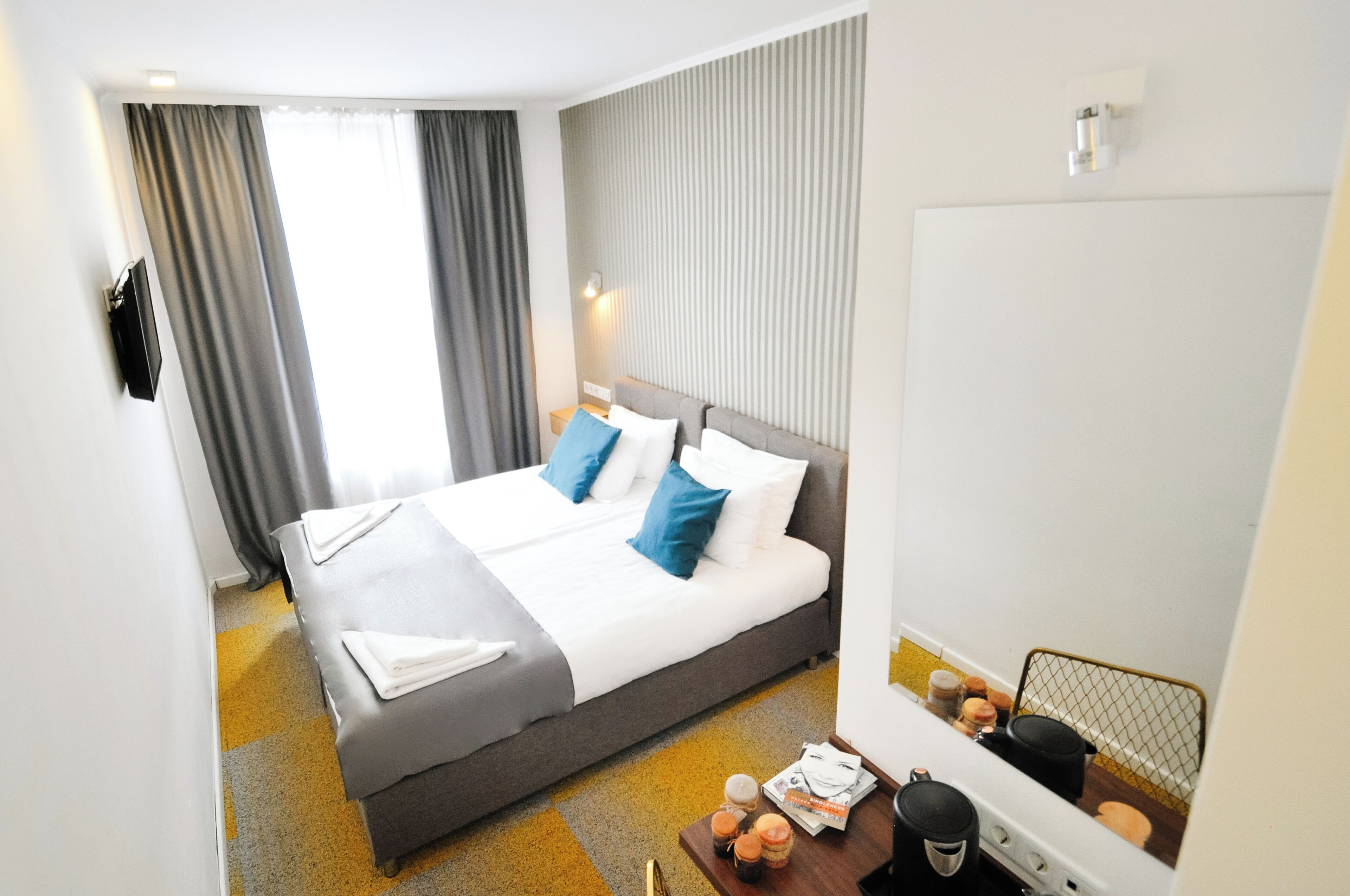A hotel room with a bed, a bedside table with a lamp, a mirror, and a window with curtains.
