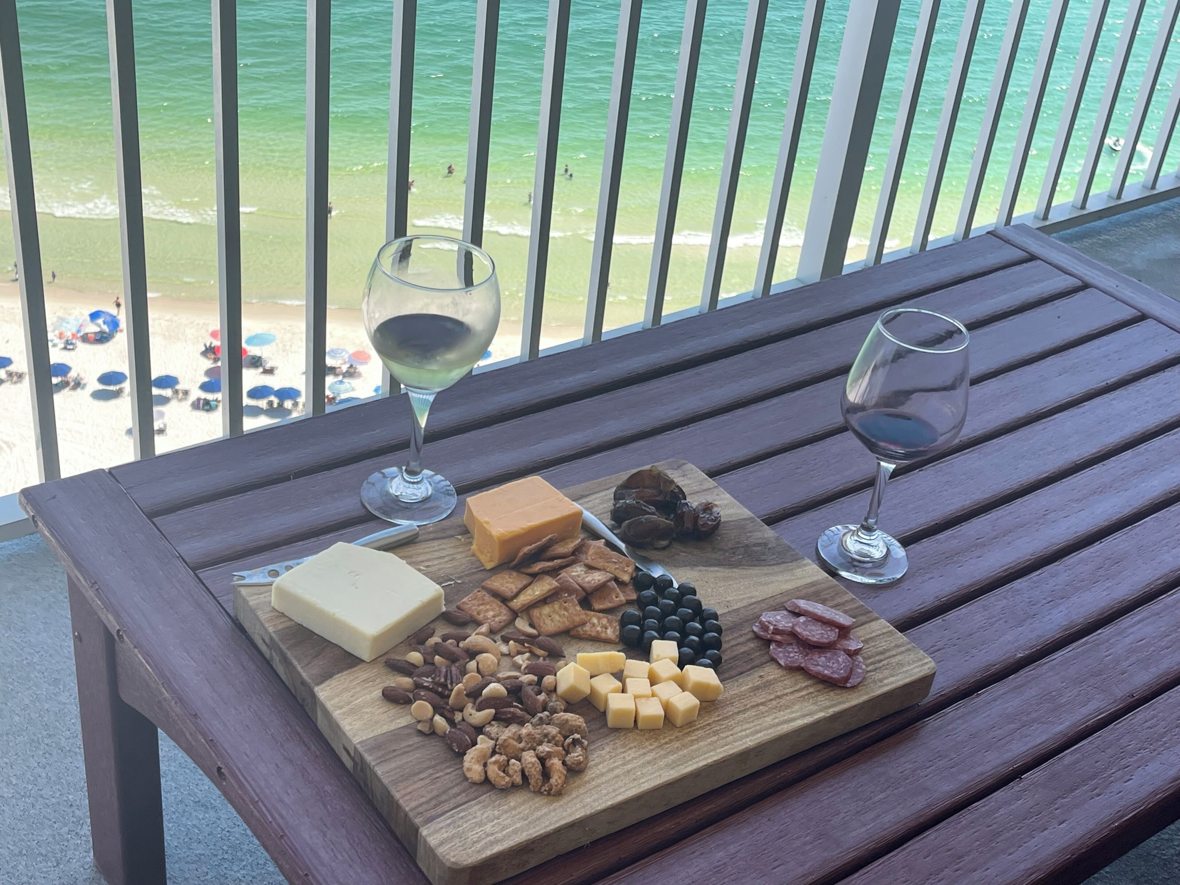 Ein Holztisch mit einer Käseplatte, Nüssen und zwei Weingläsern auf einem Balkon mit Blick auf einen Strand.