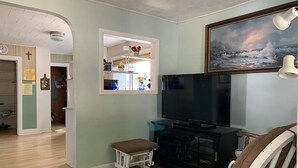 A living room with a television, a small table, and a chair. There is a painting on the wall and a door leading to another room. - Cozy Lakefront Cottage (Winchendon)