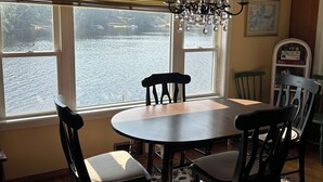 A dining area with a round table, four chairs, a chandelier, and a view of a body of water through large windows. - Cozy Lakefront Cottage (Winchendon)