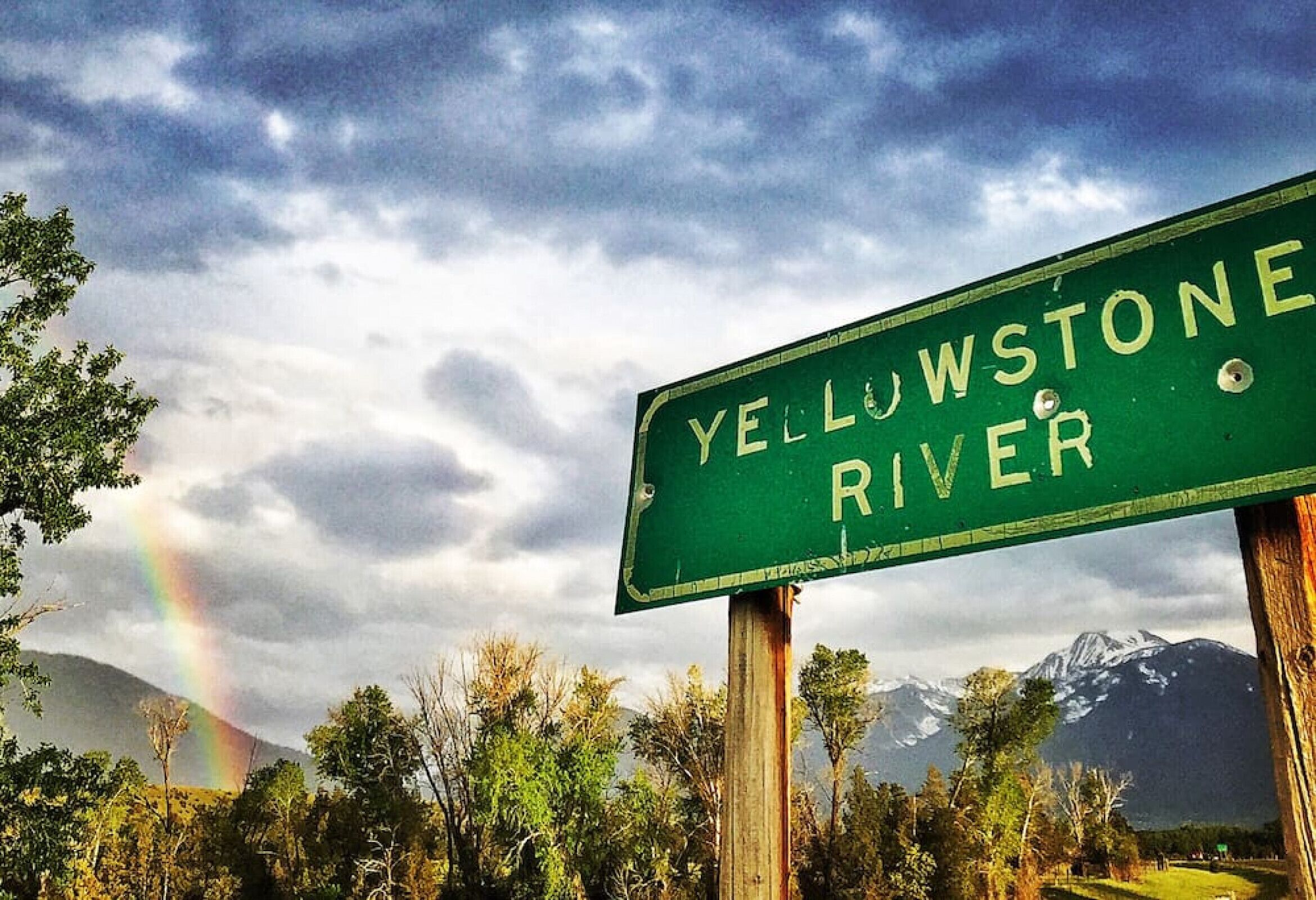 Un panneau vert sur lequel on peut lire « YELLOWSTONE RIVER », le tout sur un fond représentant un arc-en-ciel, des arbres et des montagnes.