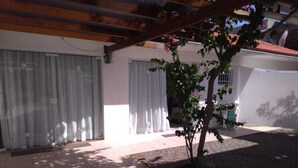 Un'area patio coperta con pavimento in piastrelle, un piccolo albero e un edificio sullo sfondo con pareti bianche e tetto in tegole rosse.