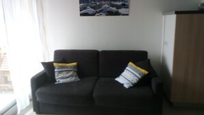 Ein Wohnzimmer mit einem dunkelgrauen Sofa, zwei gemusterten Kissen, einer Wanduhr und einem gerahmten Bild einer verschneiten Landschaft.