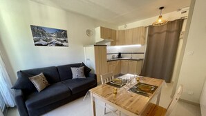 Ein kompakter Wohnbereich mit Esstisch für vier Personen, einem dunkelgrauen Sofa mit Zierkissen, einer Küchenzeile mit Holzschränken und einem an der Wand hängenden Bild von einer verschneiten Landschaft.