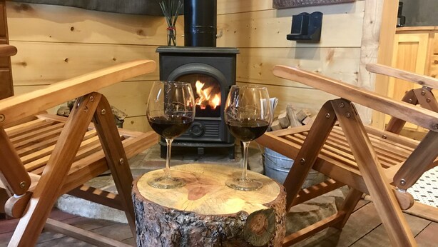 A cozy cabin interior with a wood-burning stove, two wooden chairs, and two wine glasses on a tree stump.