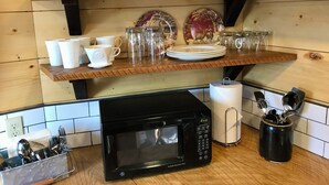 A kitchen with wooden shelves holding white cups, plates, and glass mugs. A black microwave oven is on the counter.