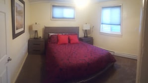 Une chambre à coucher comprenant un lit, deux tables de chevet, deux lampes, un tableau encadré au mur et deux fenêtres équipées de stores.