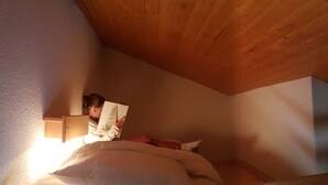 Ein kleiner, gemütlicher Raum mit Holzboden und -wänden, einem Einzelbett mit weißer Matratze und roter Bettwäsche sowie einer Nachttischlampe.