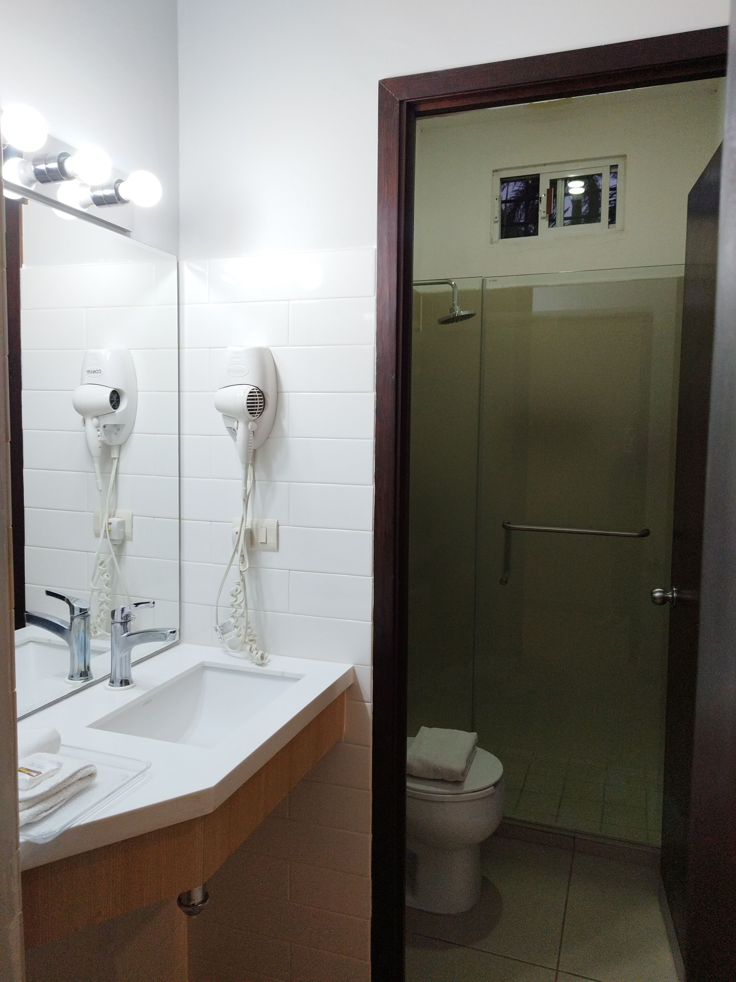 A bathroom with a mirror, two hair dryers, a sink, a toilet, and a glass-enclosed shower.