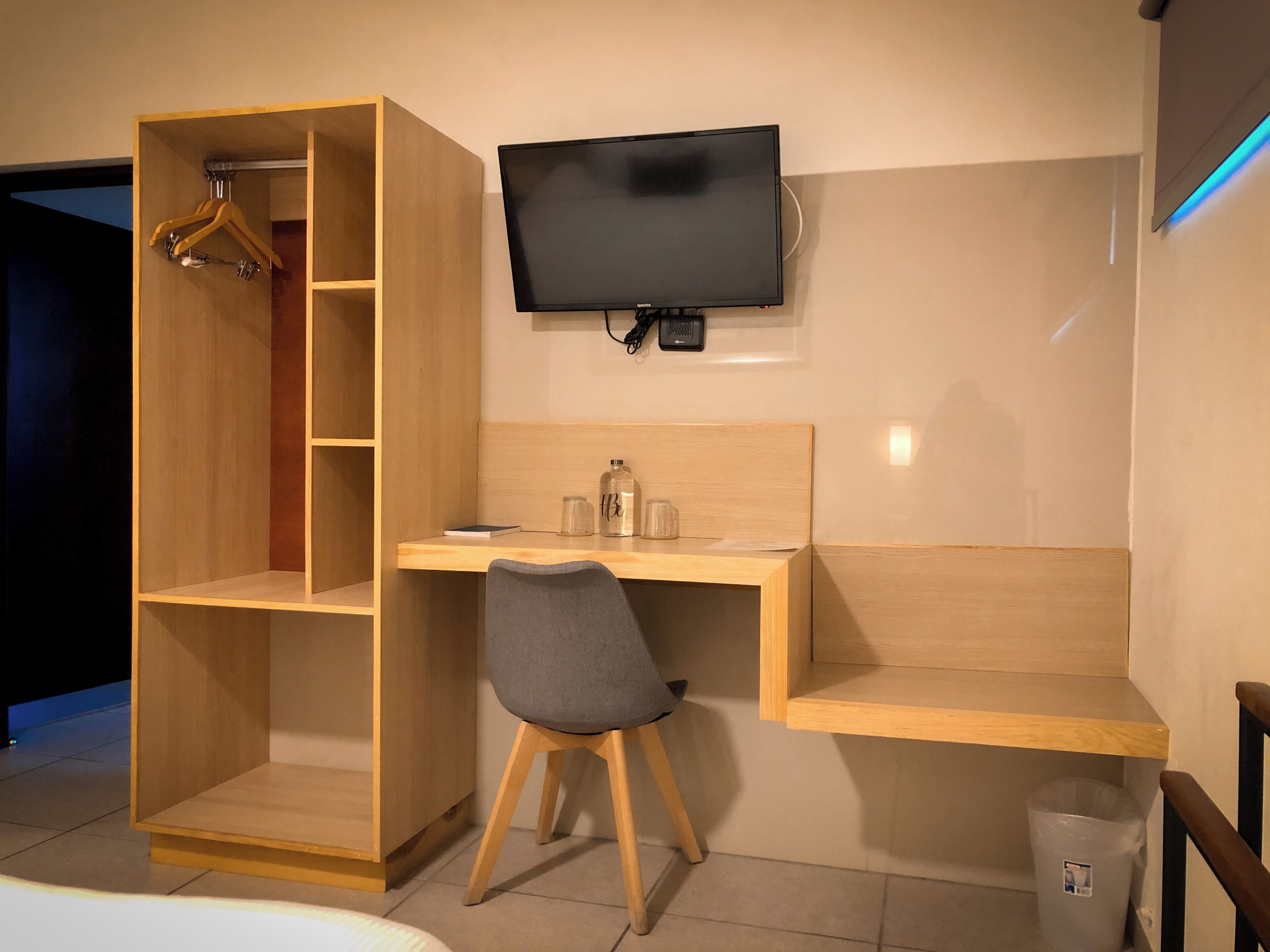 A room with a wooden desk, a chair, a TV mounted on the wall, a wardrobe with hanging space, and a trash bin.