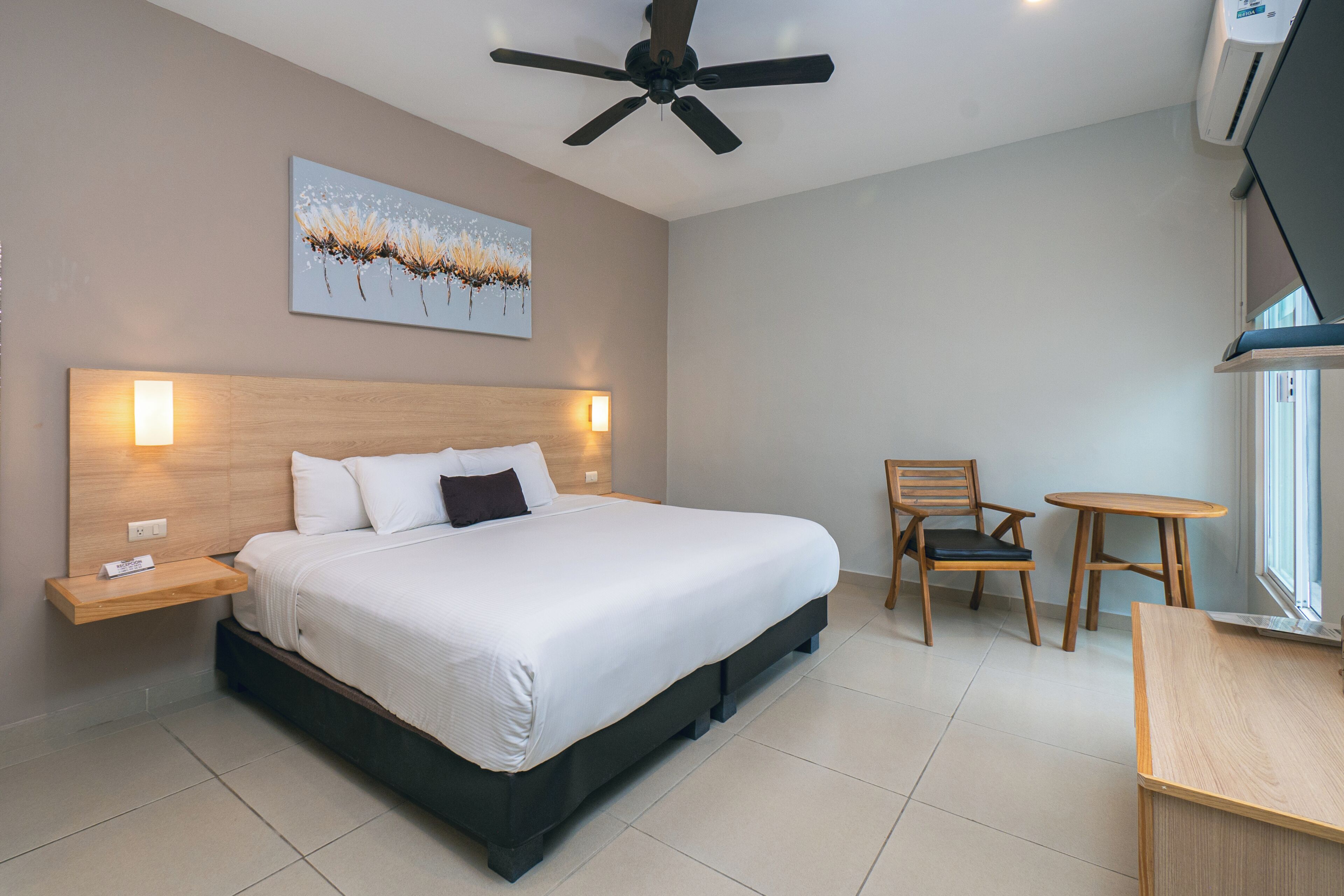 Habitación de hotel con cama, mesitas de noche, ventilador de techo, un cuadro, una silla, una pequeña mesa y una ventana.