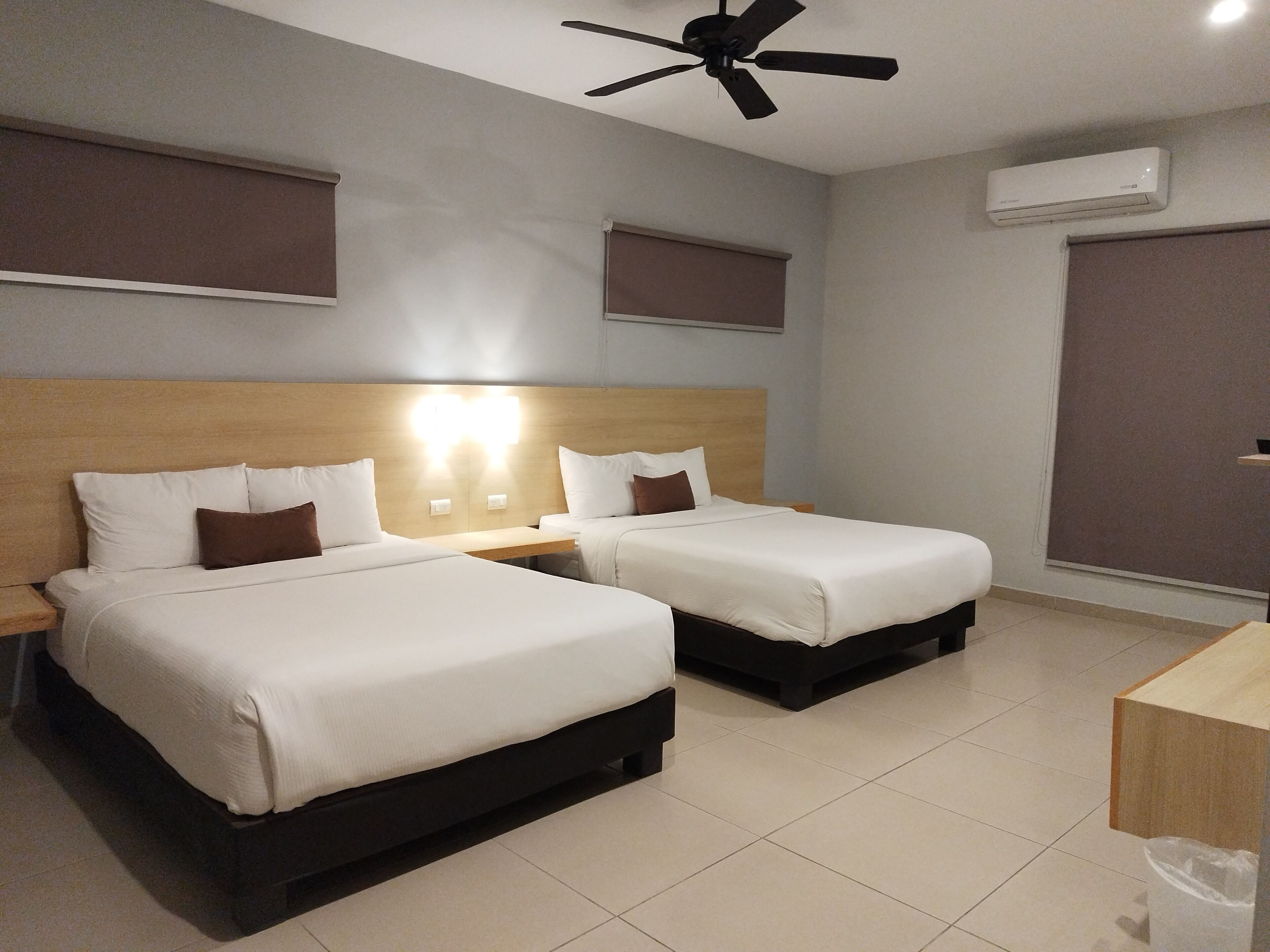 A hotel room with two beds, a ceiling fan, an air conditioning unit, and a bedside table.