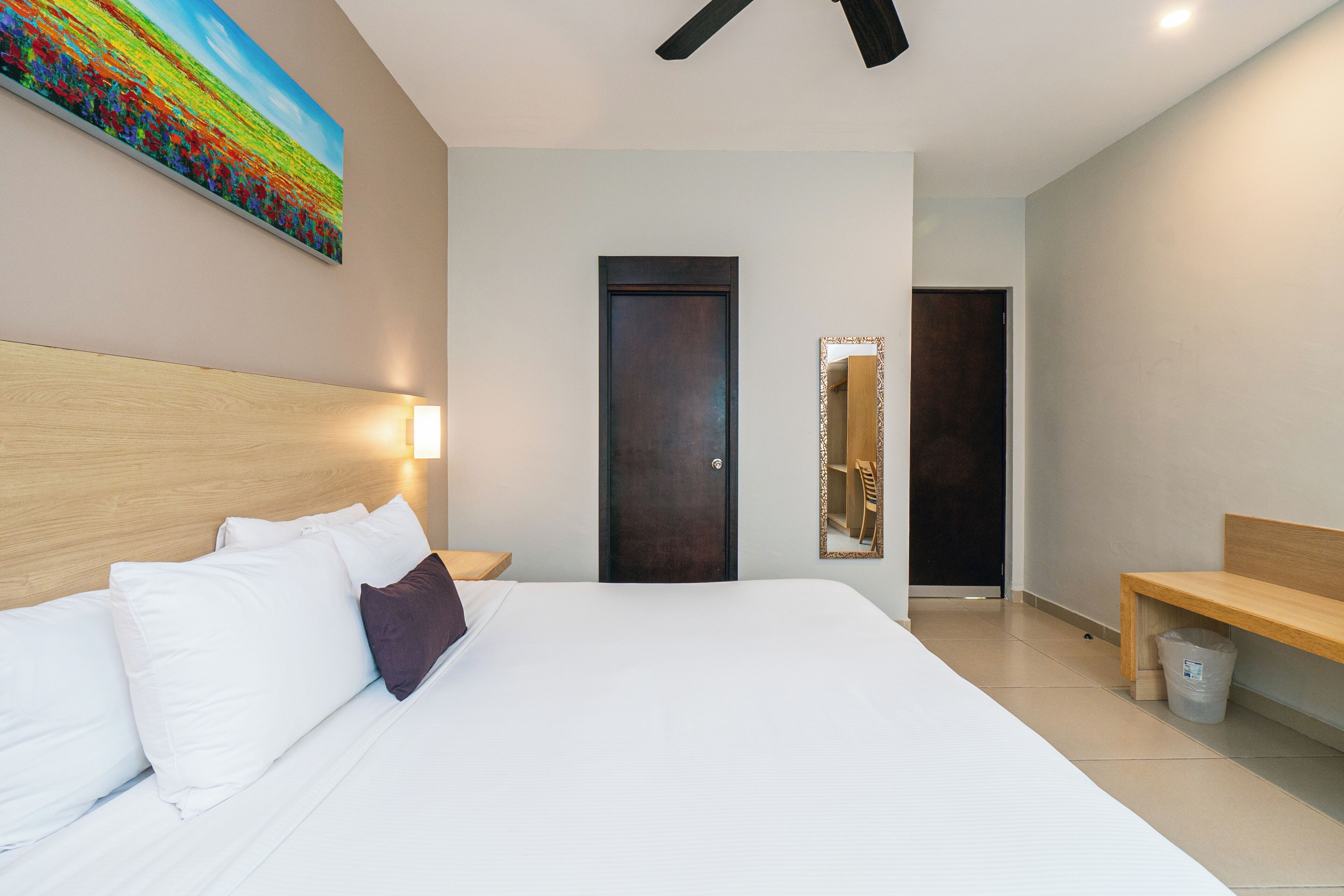 Habitación de hotel con una cama grande, cabecera de madera, un cuadro en la pared, una puerta, un espejo, una banca y un cubo de basura.