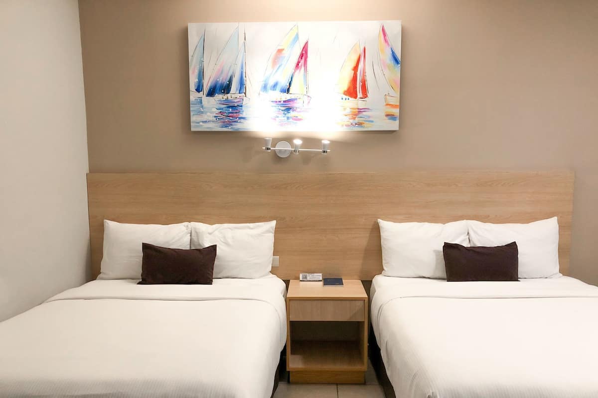 A hotel room with two beds, a wooden headboard, a nightstand with a lamp, and a painting of sailboats on the wall.