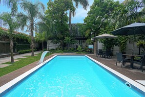 A rectangular swimming pool with a clear blue water surface, surrounded by a well-maintained garden with various trees and shrubs. - Biza Express (Ciudad Valles)