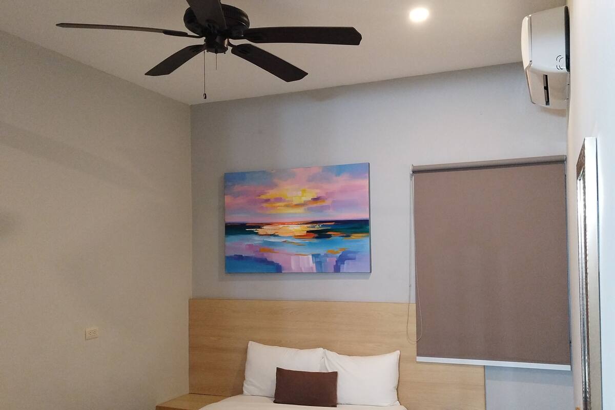 A hotel room with a bed, a ceiling fan, a painting, a window, and a wall-mounted air conditioner.