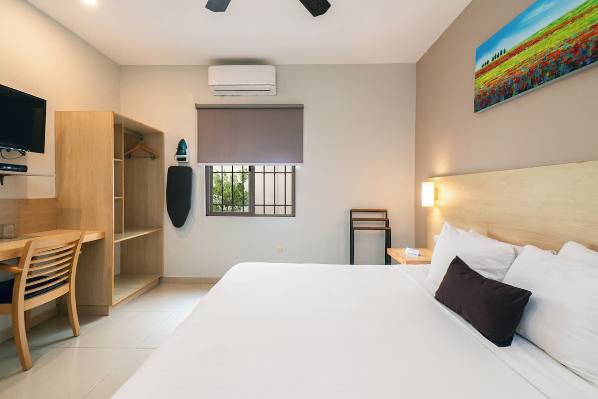 A hotel room with a bed, a desk, a chair, a wardrobe, a window, and an air conditioning unit.