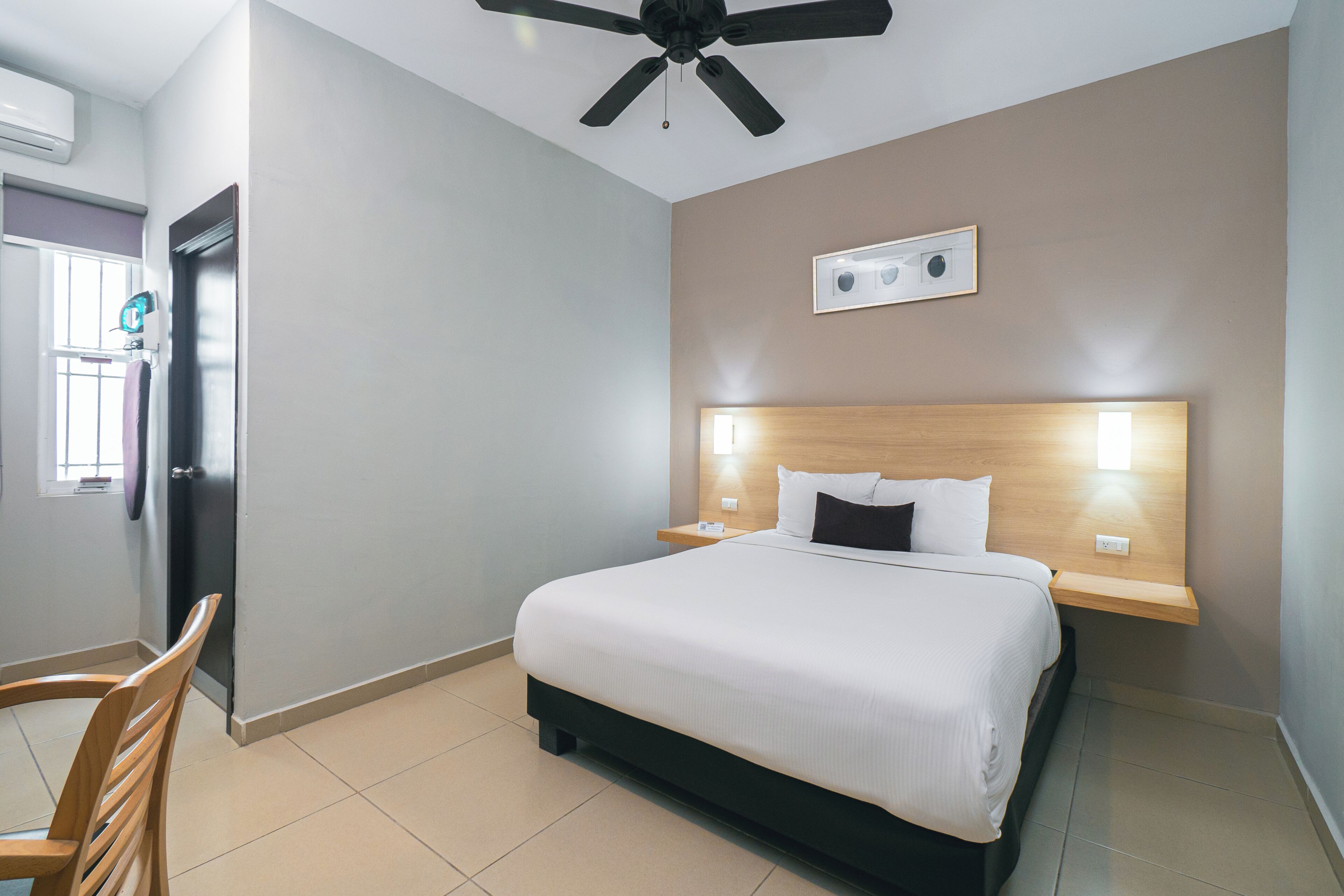 A hotel room with a bed, a ceiling fan, a bedside table, and a wall-mounted light.