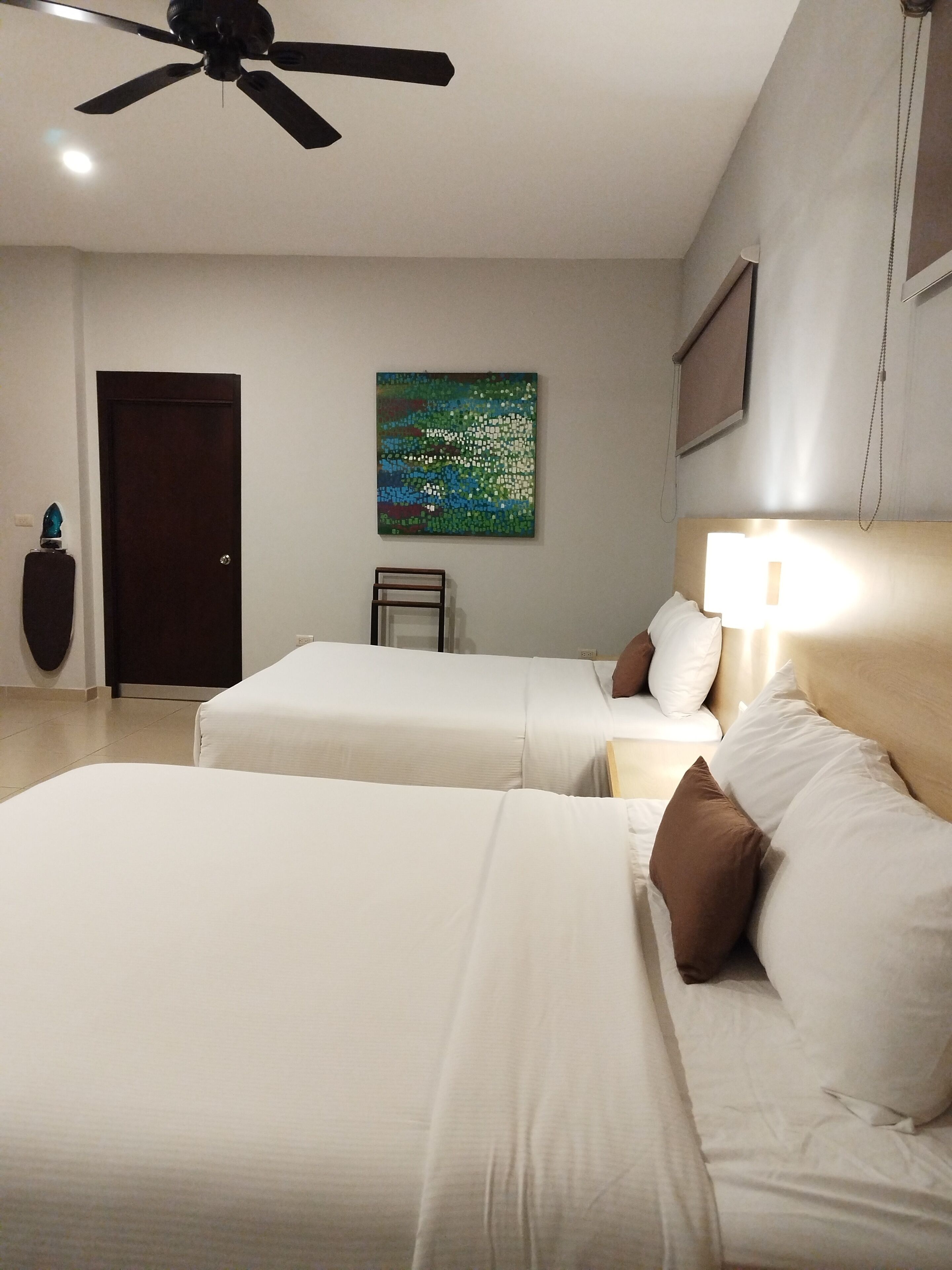 A hotel room with two beds, a ceiling fan, a painting on the wall, a door, and a bedside table.
