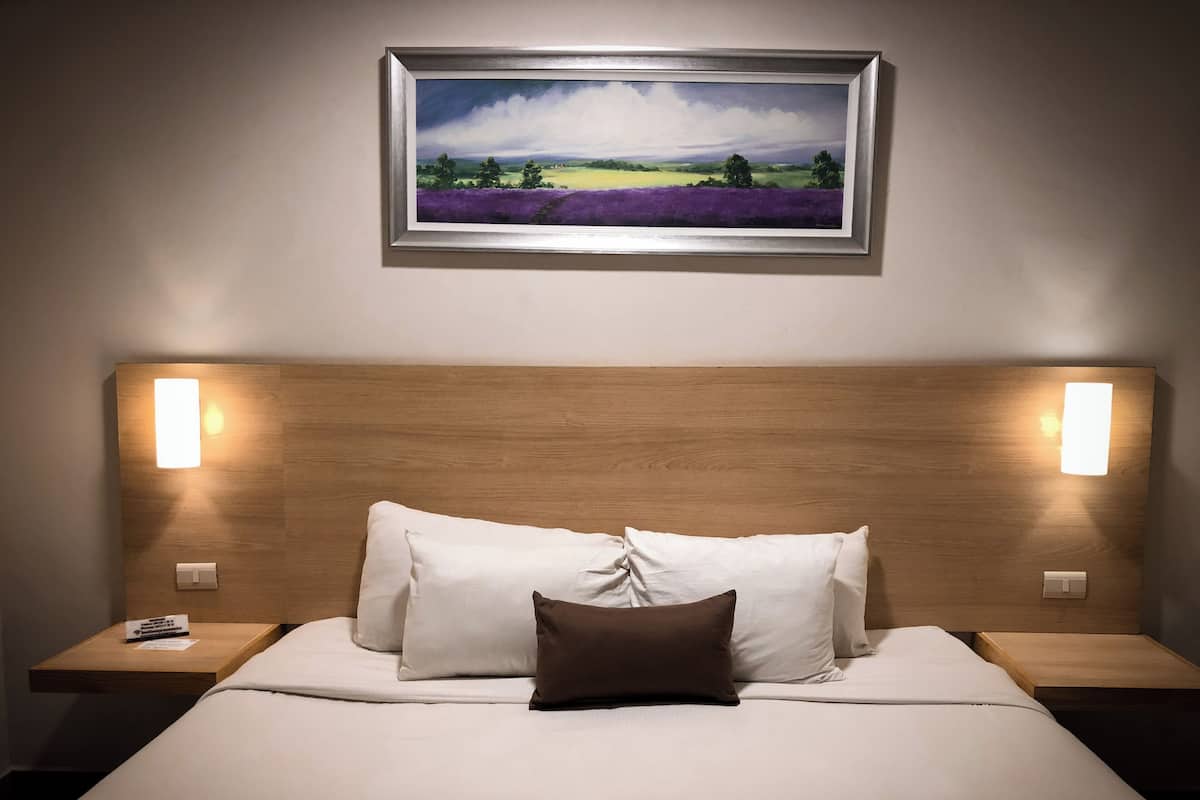 A hotel room with a wooden headboard, white bedding, a brown pillow, and a framed landscape painting above the bed.