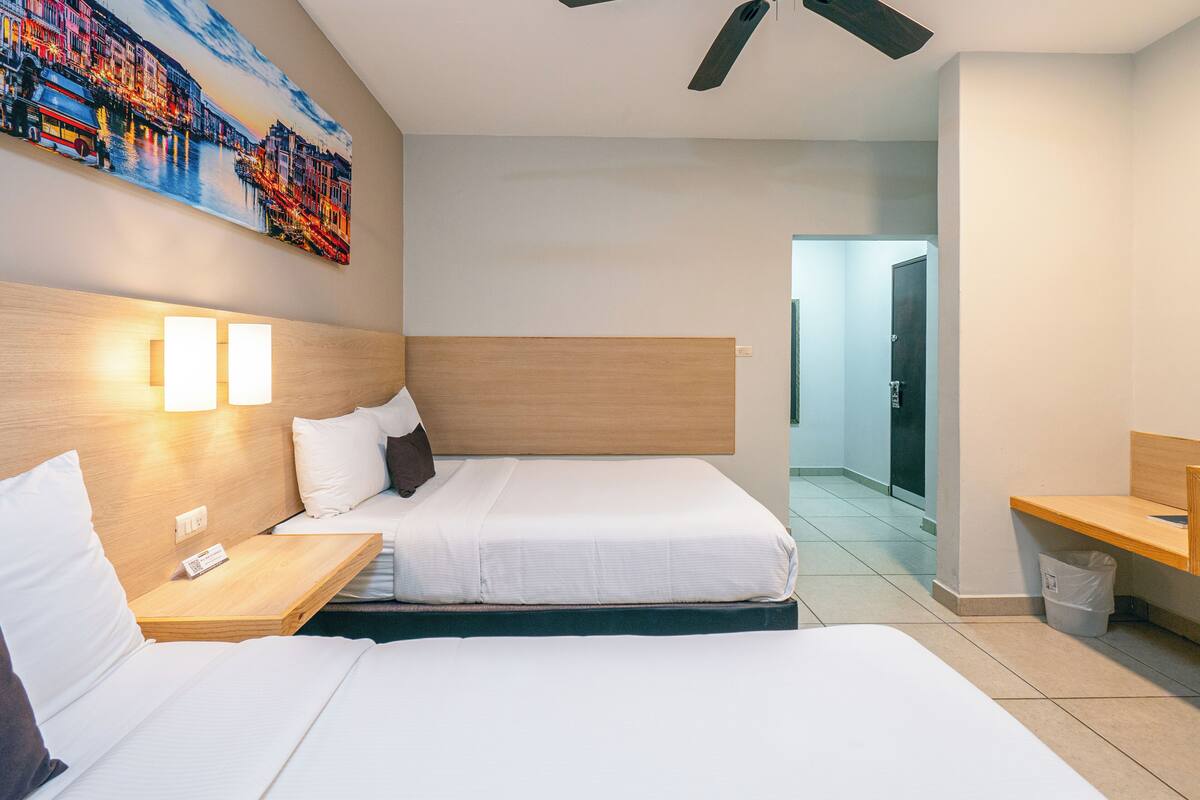 A hotel room with two beds, a wooden headboard, bedside tables with lamps, a painting on the wall, and a view of the hallway.