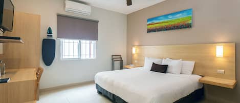 A hotel room with a bed, a desk, a chair, a ceiling fan, an air conditioner, a window with blinds, a painting on the wall, and a nightstand with a lamp.