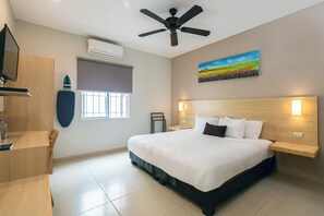 A hotel room with a bed, a desk, a chair, a ceiling fan, an air conditioner, a window with blinds, a painting on the wall, and a nightstand with a lamp. - Biza Express (Ciudad Valles)