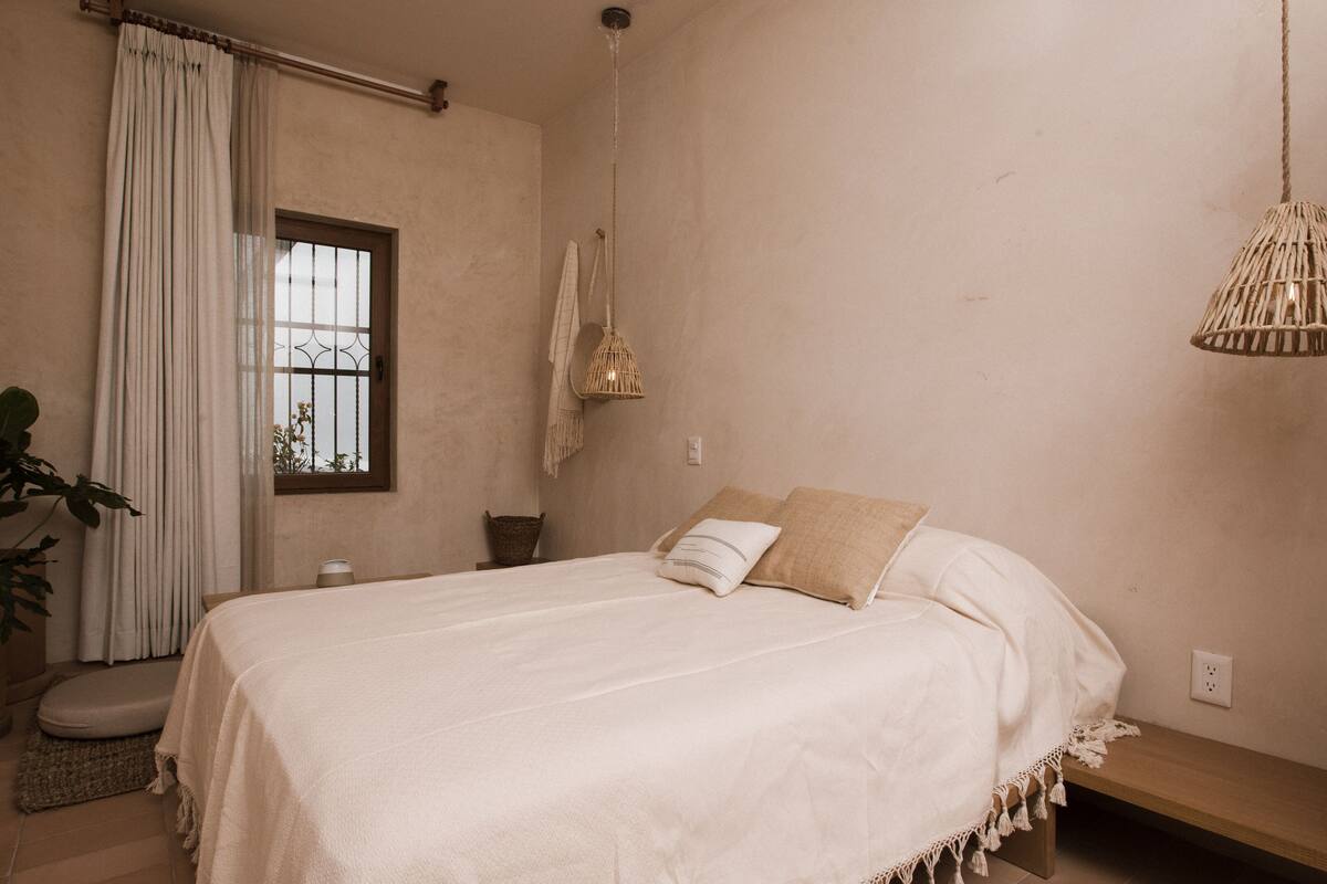 Ein Schlafzimmer mit Bett, Nachttisch, Pendelleuchte, Fenster mit Vorhängen und einer Topfpflanze.