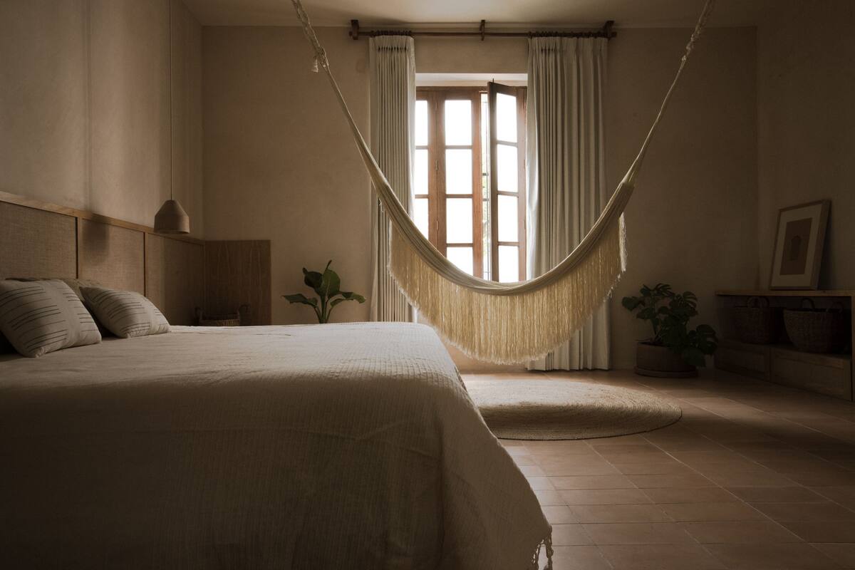 Ein Schlafzimmer mit Hängematte, Bett, Nachttisch, Lampe und einem Fenster mit Vorhängen.