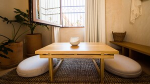 A room with a wooden table, two white cushions, a potted plant, a woven basket, and a view through a window with a metal gate. - Casa Montore by Barrio Mexico (Guadalajara)