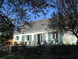 A two-story house with a sloped roof, green shutters, and a small balcony with a table and chairs.