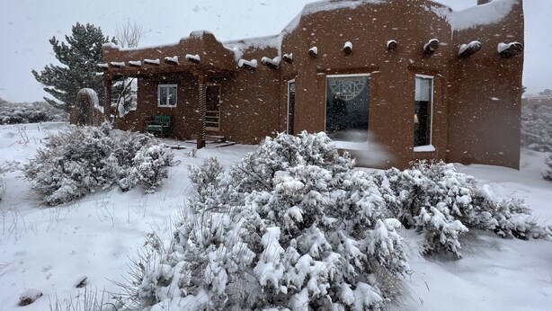Una casa de adobe rústica con exterior nevado, un porche de madera y un ventanal con diseño decorativo.