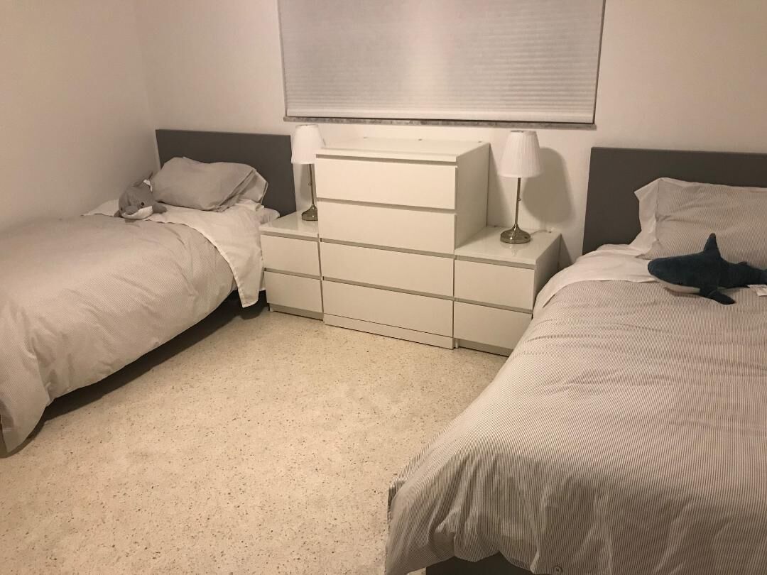 Ein Doppelzimmer mit zwei Betten, jeweils mit einem Kopfkissen und einer Lampe. In der Mitte des Zimmers befindet sich eine Kommode mit Schubladen.