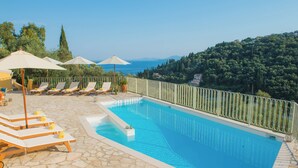 A swimming pool with lounge chairs and umbrellas, surrounded by a stone deck and a metal railing, overlooking a hillside. - T' adelfia  -  ERMIS (Corfu)
