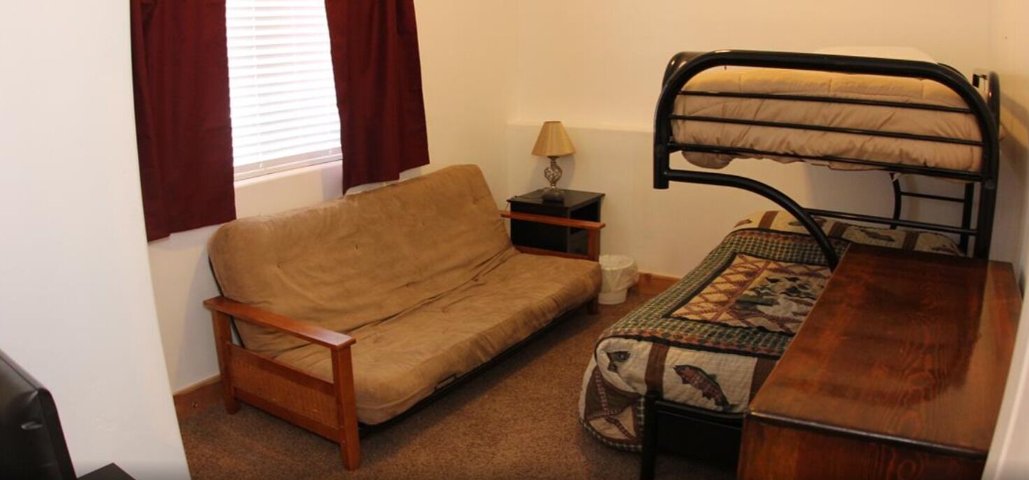 A compact room with a wooden sofa, a bunk bed, a small bedside table with a lamp, and a patterned quilt.