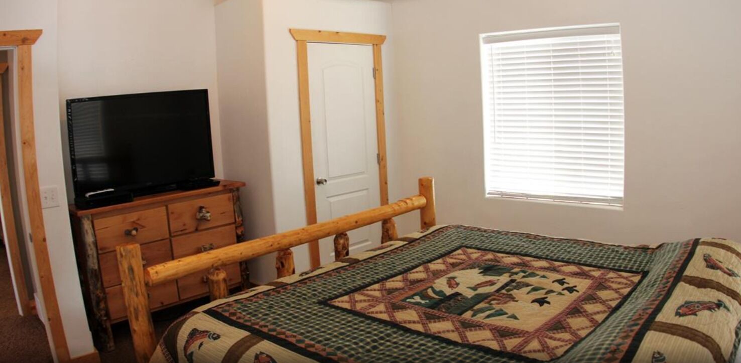 A bedroom with a wooden bed frame, a television on a dresser, a door, and a window with blinds.
