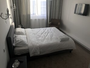 Una cama bien hecha con sábanas blancas, una mesita de noche con un control remoto y recipientes de vidrio, una silla y un televisor de pantalla plana montado en la pared.