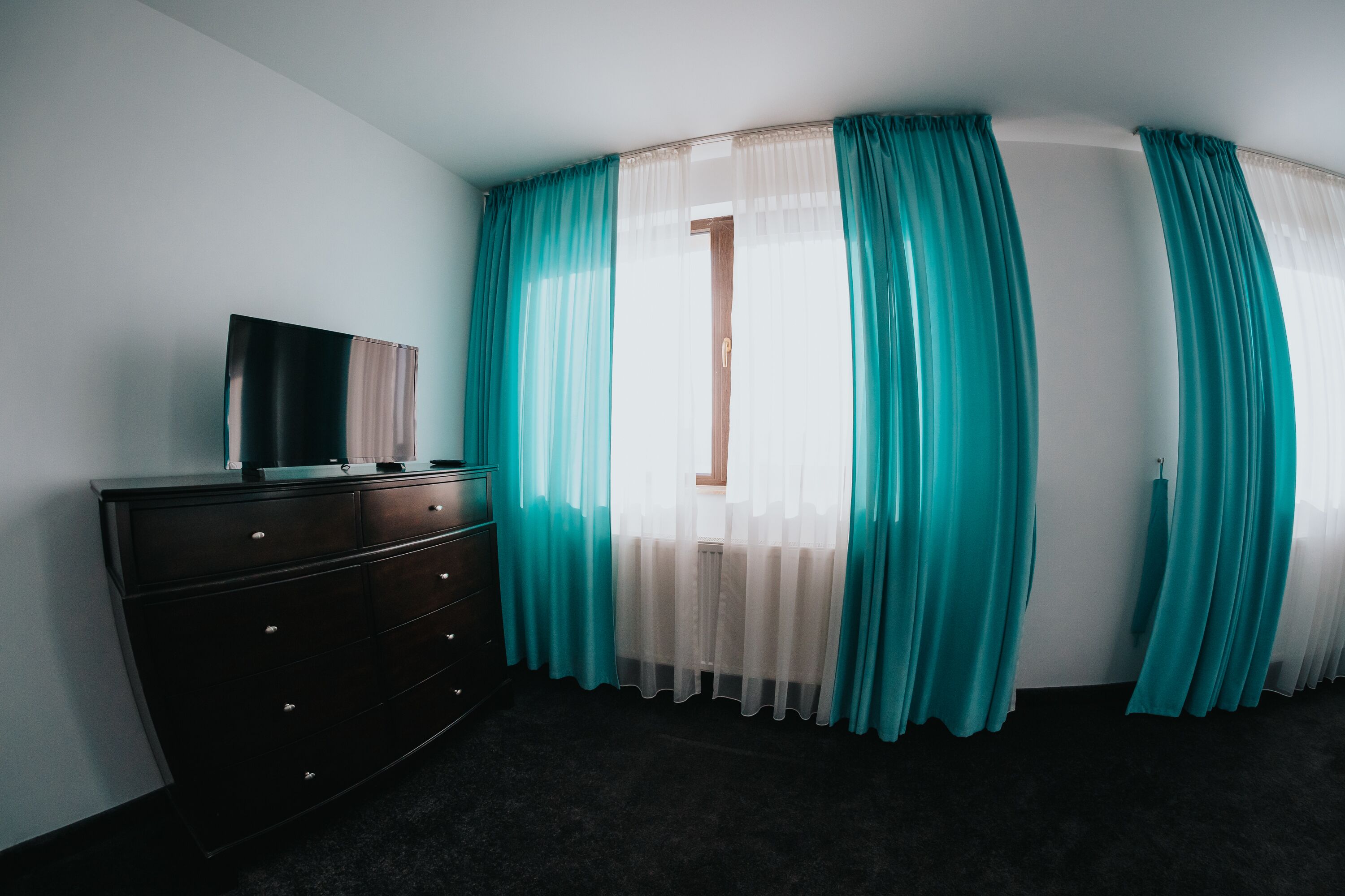 A room with a dark wooden dresser, a television on top, teal curtains, and a window with a view.
