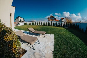 Un jardín trasero con un patio de piedra, dos sillones de descanso, césped y una fila de árboles.