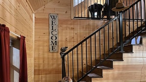 Un intérieur en bois avec un escalier, une pancarte indiquant « BIENVENUE » et un chapeau posé sur un objet décoratif.