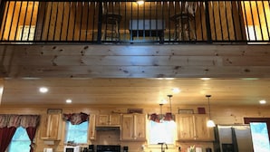 L’intérieur d’un chalet en bois comprend un coin cuisine, un espace repas avec une table et des chaises, ainsi qu’un balcon doté d’une balustrade.