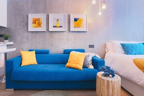 Una sala de estar moderna con un sofá azul, cojines amarillos y una mesita redonda de madera. Encima del sofá hay tres cuadros enmarcados en la pared.