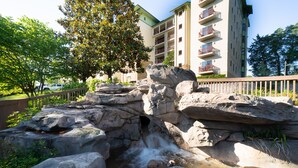 A natural rock formation with a small waterfall, surrounded by greenery and a multi-story building with balconies in the background.